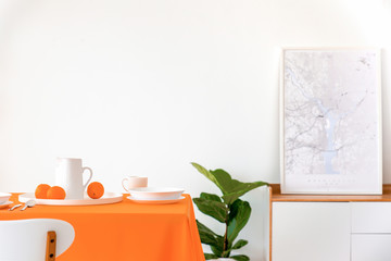 Poster on cabinet next to plant and table with orange cloth in white dining room interior. Real photo