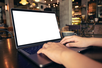 Mockup image of hands using and typing at laptop with blank white desktop screen with coffee cup on wooden table in cafe