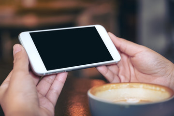 Mockup image of hands holding and using a white mobile phone with blank black screen horizontally for watching with coffee cup on wooden table