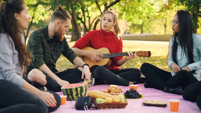 Romantic young people are singing and playing the guitar on picnic sitting on plaid on lawn in park and having fun. Musical instruments and culture concept.