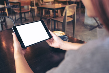 Mockup image of woman's hands holding black tablet pc with blank white screen with coffee cup on wooden table in cafe