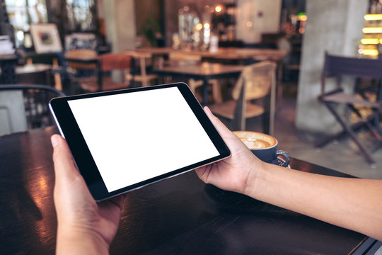Mockup image of hands holding black tablet pc with blank white screen with coffee cup on wooden table in cafe