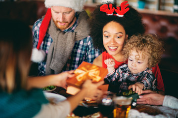 Man and mixed race woman giving toddler  Christmas gift while sitting at table. Christmas holidays concept.