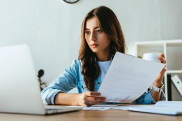 portrait of concentrated businesswoman with cup of coffee and papers at workplace with  laptop in office
