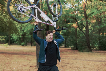 Young cuacasian man crashes the bicycle hodlig it up in both hands, ahgry expression on the face. Outdoors, park around him.