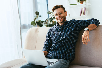 portrait of smiling man in eyeglasses with laptop sitting on sofa at home