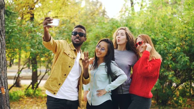 African American student is taking selfie with beautiful girls Asian and Caucasian standing in park, using smartphone and posing for camera. Youth and photo concept.