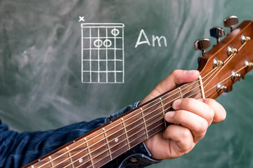 Man in a blue denim shirt playing guitar chords displayed on a blackboard, Chord A minor