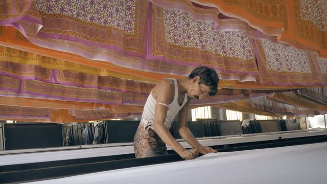 A movement shot of a daily wage worker or laborer working in textile printing interior factory setup while colorful printed cotton sheets hanged on the top to let them dry