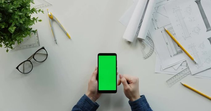 Close up of the white desk with drawings equipment and graphics of the female worker and Caucasian hands using black vertical smartphone with green screen. Top view.