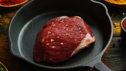 From above shot of piece of fresh uncooked meat with spices lying on frying pan on timber tabletop