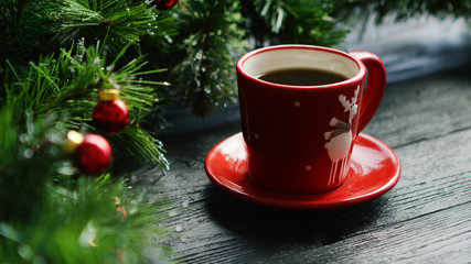Obraz na płótnie Canvas Closeup shot of small cup of fresh hot drink standing on lumber tabletop near fresh conifer branches decorated with small red beads