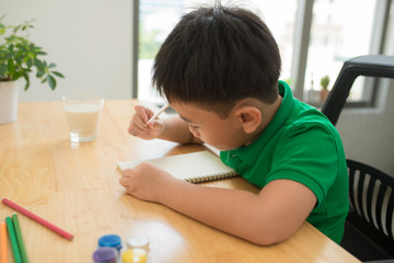 Cute confused smiling boy doing homework, coloring pages, writing and painting . Children paint. Kids draw. Preschooler with books in the library. Colorful pencils and paper on a desk. Creative boy.