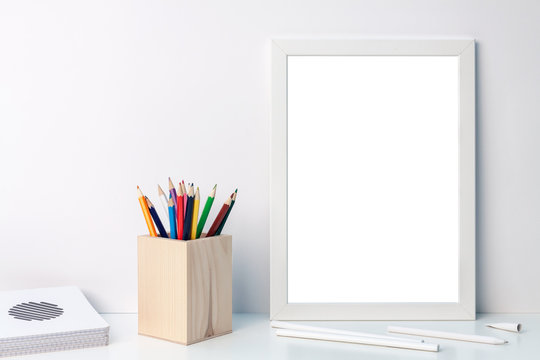 Photo of a white mockup frame with colored pencils in a wooden toolbox with notebooks on the desk