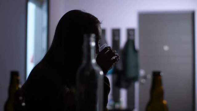Silhouette of wasted drunk woman with alcohol addiction sitting alone in dimmed room and drinking alcohol beverage. Many empty bottles on foreground. Dolly shot.