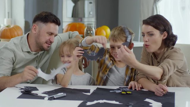 Joyous little boy and girl sitting with parents at table in the living room and playing with paper bats while making Halloween decorations