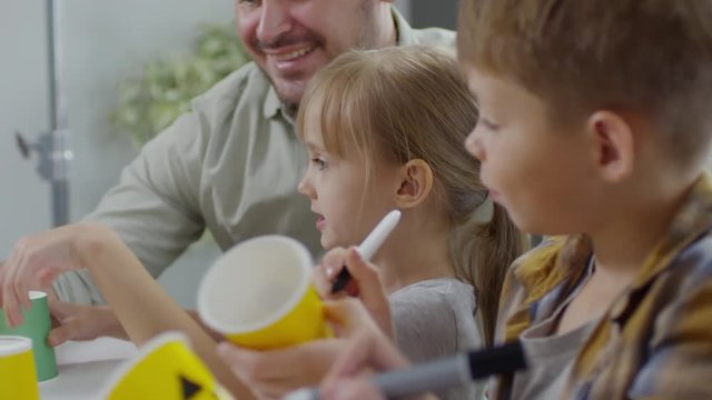 Close-up of little boy and girl drawing on paper cup and talking with father while making decorations for Halloween party