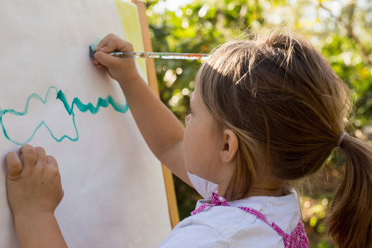 A pre-school girl painting at an easel in a garden. 
