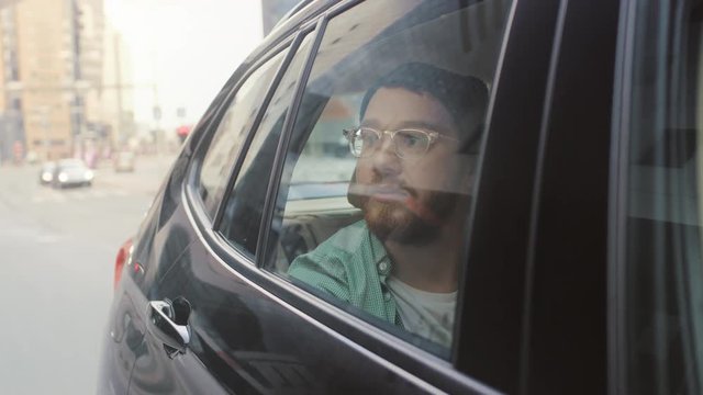 Stylish Young Man Rides on a Passenger Back Seat of a Car, Looks out of the Window in Wonder. Big City View Reflects in the Window. Camera Mounted outside Moving Car. 