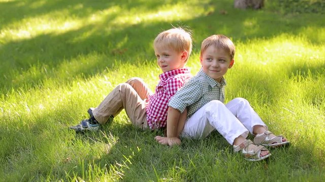 Two cheerful little boys of 4 years old are sitting back to back in the park on the grass. Two young children relax in the summer in nature.