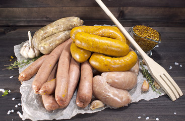 Assorted raw sausages with spices, vegetables and mustard on crumpled paper on a wooden table.