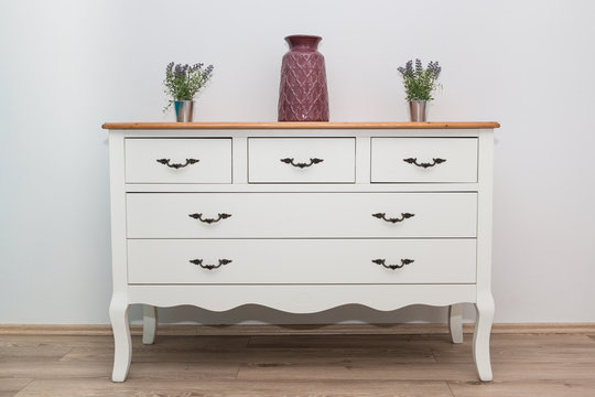 White wooden dresser with three vases and flowers on white wall background. Chest of drawers close up.