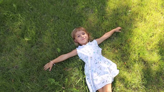 Little cute girl in white dress lying on green grass in Sunny Park in hot weather. Unusual little girl with different eye color in the Park, portrait. The view from the top.