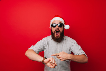 Amazed bearded man in santa claus hat pointing at smartwatch over red background