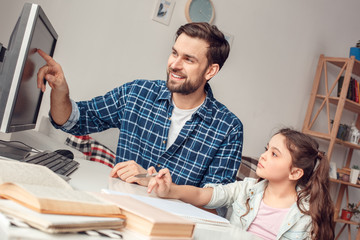 Father and little daughter at home sitting at table man pointing at monitor explaining information...