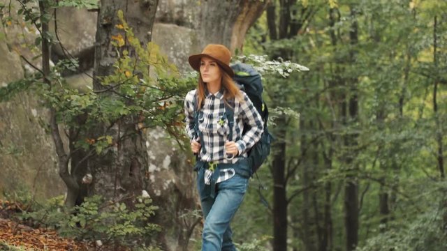 Slim, good-looking woman, in elegant brown hat and stylish casual clothes, backpacking through mountains, outdoor shot in beautiful rocky countryside among green, tall trees