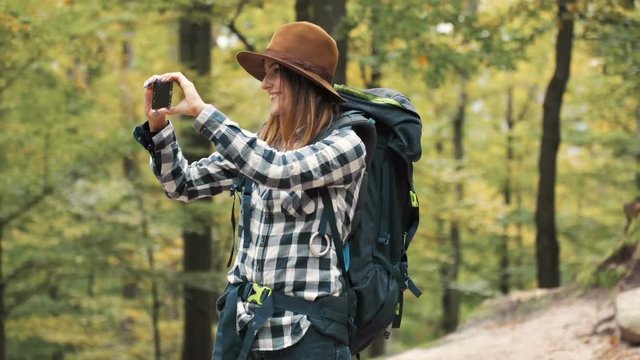 Happy, female backpacker taking photo with smartphone on amazing hiking trip, standing cheerfully in lovely, fall forest