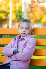 A girl in a pink coat folded her arms in front and looking at the camera, sitting on a yellow bench in the park.