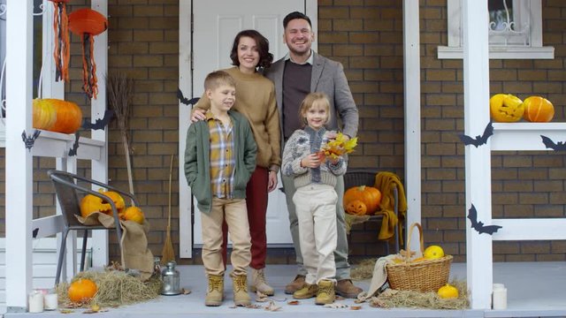 Joyous mother and father standing with kids on the porch decorated for Halloween with jack-o-lanterns, bats and brooms, happily smiling and looking at camera
