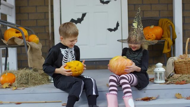 Happy little brother and sister dressed like witch and skeleton sitting on porch decorated for Halloween, playing with jack-o-lanterns and laughing