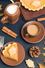 Piece of pumpkin pie with cinnamon and coffee with milk in brown dishes on black background with autumn yellow leaves.