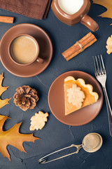Piece of pumpkin pie with cinnamon and coffee with milk in brown dishes on black background with autumn yellow leaves.