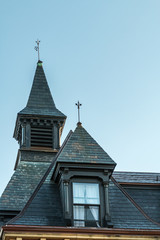 An older house with a dormer window and cupola. There are two roofs that are steep with tiles. Each roof has a lighting rod. There is a blue sky in the background.