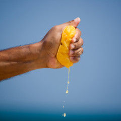 Close up of hand squeezing orange, citrus with tropical sea backdrop, and blue sky