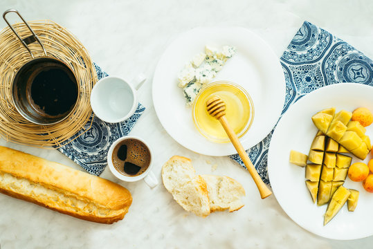  couple of cups with coffee, cheese, honey, baguette and fruit on a light marble table