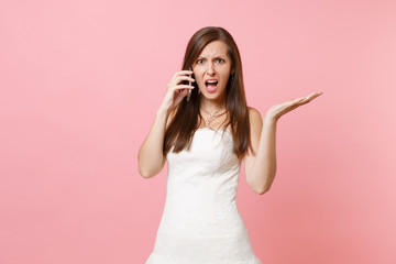 Indignant bride woman in white wedding dress have problem with planning wedding talking on mobile phone, swearing isolated on pastel pink background. Wedding to do list. Organization of celebration.