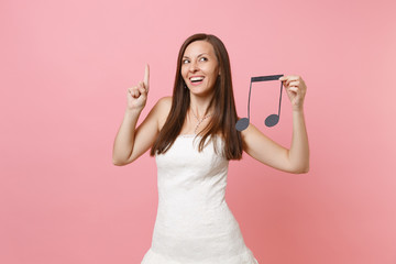 Dreamy bride woman in wedding dress pointing index finger up hold musical note choosing staff, musicians or DJ isolated on pink background. Wedding to do list. Organization of celebration. Copy space.