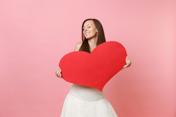 Tender happy bride woman with closed eyes in elegant white wedding dress holding empty blank red heart isolated on pastel pink background. Wedding celebration concept. Copy space for advertisement.