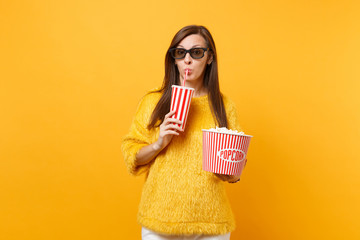 Pretty young woman in 3d imax glasses watching movie film holding bucket of popcorn, drinking cola or soda from plastic cup isolated on yellow background. People sincere emotions in cinema, lifestyle.