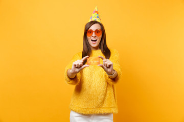 Shocked amazed young woman in orange heart glasses and birthday party hat holding credit card, enjoying holiday, celebrating isolated on yellow background. People sincere emotions, lifestyle concept.