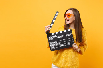 Laughing young woman in orange heart eyeglasses looking aside and holding classic black film making...