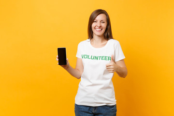 Woman in white t-shirt written inscription green title volunteer hold mobile phone with blank empty screen isolated on yellow background. Voluntary free assistance help, charity grace work concept.
