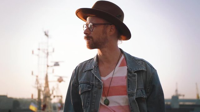 Portrait of young stylish guy with red beard in glasses and in hat looking at camera during sunset outdoors. Cinematic shot