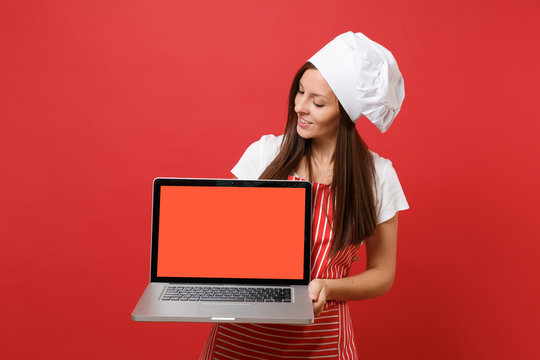 Housewife female chef cook or baker in striped apron white t-shirt toque chefs hat isolated on red wall background. Woman hold laptop pc blank screen for promotional content Mock up copy space concept