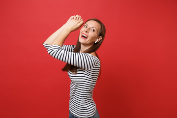 Portrait of joyful young girl in striped clothes with wireless earphones dancing rising hands listening music isolated on red background. People sincere emotions lifestyle concept. Mock up copy space.