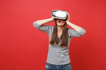 Amazed young girl in striped clothes, virtual reality glasses keeping mouth wide open, putting hands on head isolated on red background. People sincere emotions, lifestyle concept. Mock up copy space.